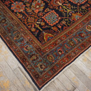 Corner view of a dark blue rug with elaborate red and teal borders on weathered wood floors.