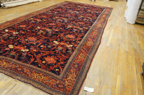 Large navy and rust-red floral patterned rug with multiple borders rests on light wooden planks.