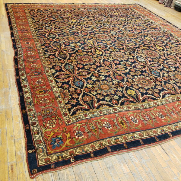 Navy blue field rug with repeating multicolored floral motifs and an elaborate rust red border on wood floors.
