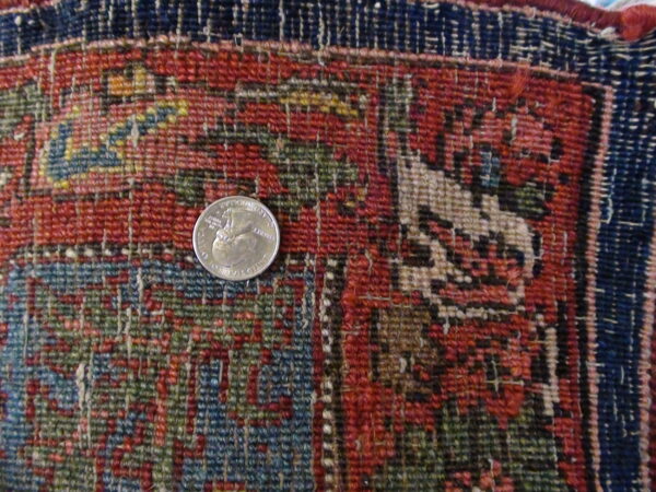 Low-pile rug segment featuring densely woven red, navy, and green geometric patterns, with a quarter for scale.