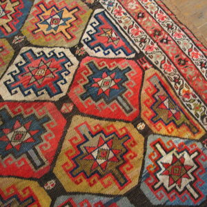 Low-pile rug with dense repeating red, blue, and gold geometric medallions rests on distressed wood flooring.