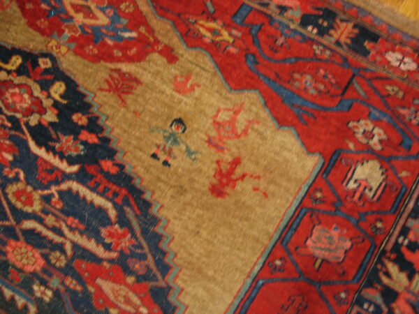 Woven rug detail showing a tan field with stylized figures bordered by red and navy geometric motifs.