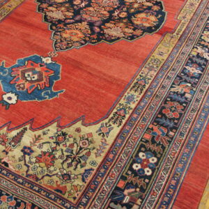 Detail view of a coral red rug featuring intricate navy, beige, and blue traditional patterning on wood flooring.