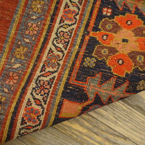 A low-pile rug with red, navy, and detailed floral-geometric patterns is rolled on wood planks.