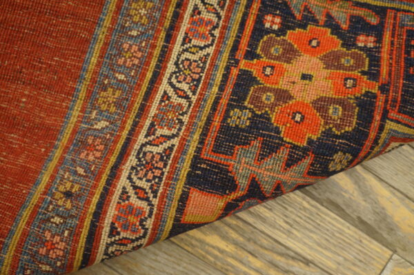 A low-pile rug with red, navy, and detailed floral-geometric patterns is rolled on wood planks.