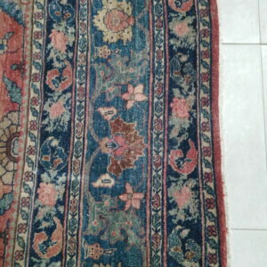 Worn traditional rug with red, blue, and tan botanical patterns rests on light square tiles.
