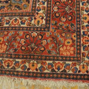 Low-pile red rug with intricate floral borders and fringe rests on yellow distressed wood planking.