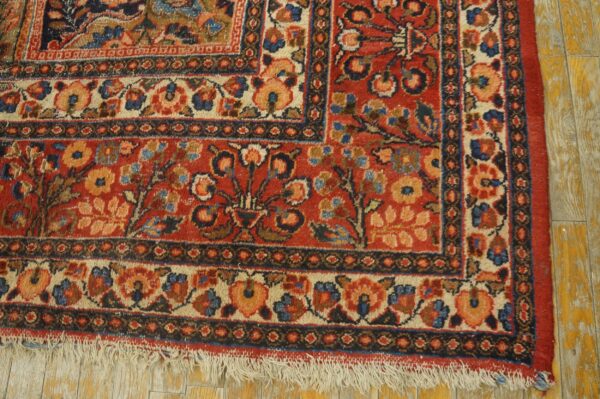 Low-pile red rug with intricate floral borders and fringe rests on yellow distressed wood planking.