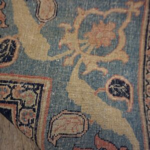 Intricate low-pile rug rolled slightly, featuring blue, tan, and pink traditional motifs over wood.