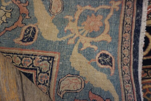 Intricate low-pile rug rolled slightly, featuring blue, tan, and pink traditional motifs over wood.