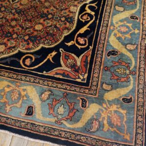 Ornate traditional rug corner in navy, rust, and teal blue sits on distressed yellow wood floor.