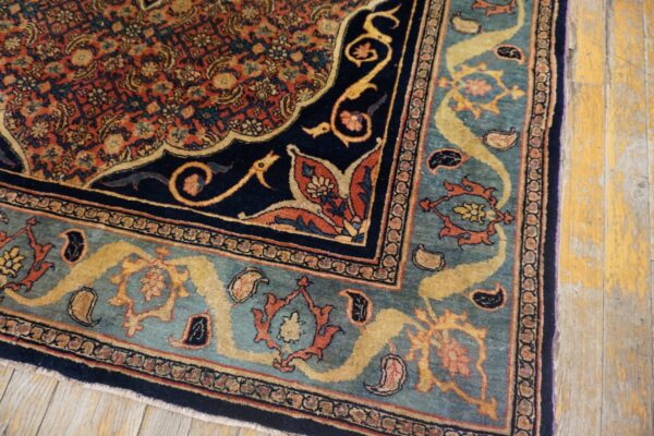 Ornate traditional rug corner in navy, rust, and teal blue sits on distressed yellow wood floor.