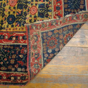 Red, blue, and golden patterned rug corner folded back, exposing the backing on wide wood planks.
