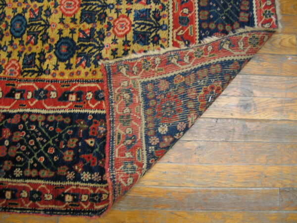 Red, blue, and golden patterned rug corner folded back, exposing the backing on wide wood planks.
