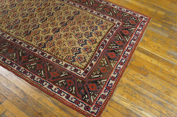 Low-pile patterned rug featuring tan, red, blue, and brown tones on worn wooden floorboards.