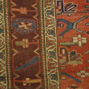 Close-up of low-pile rug showing red, brown, and blue geometric borders on light wood floorboards.