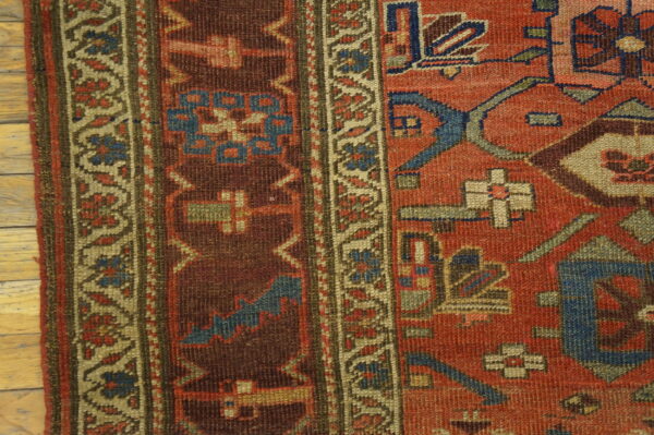 Close-up of low-pile rug showing red, brown, and blue geometric borders on light wood floorboards.