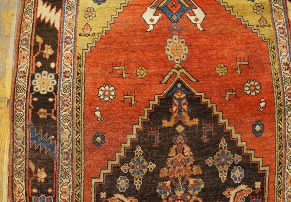 Geometric rug detail showing a rusty red field, dark stepped medallion, and stylized motifs on wooden flooring.