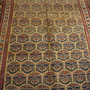 The rug displays structured rows of blue and red motifs on a gold field, with dark brown multi-striped borders.