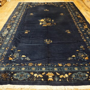 Navy area rug with intricate gold and teal floral borders displayed on wide plank wooden flooring.