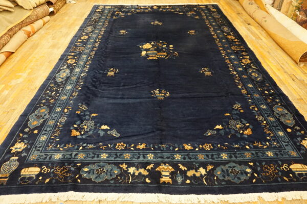 Navy area rug with intricate gold and teal floral borders displayed on wide plank wooden flooring.