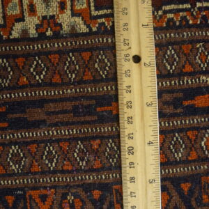 Geometric patterned rug texture in rich red, dark brown, and orange tones, measured by a vertical wooden ruler.