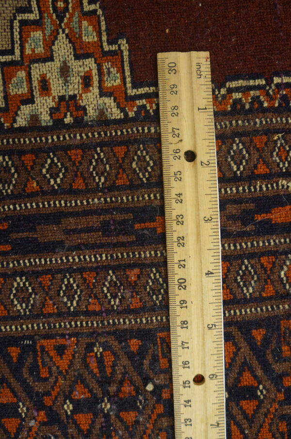 Geometric patterned rug texture in rich red, dark brown, and orange tones, measured by a vertical wooden ruler.