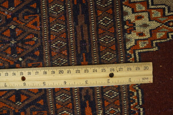 A wooden ruler rests on a low-pile geometric rug featuring dark brown, rust, and cream colors.