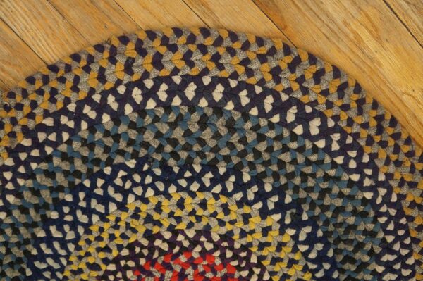 Rounded, braided rug in blue, yellow, gray, and red colors rests on light wooden floorboards.