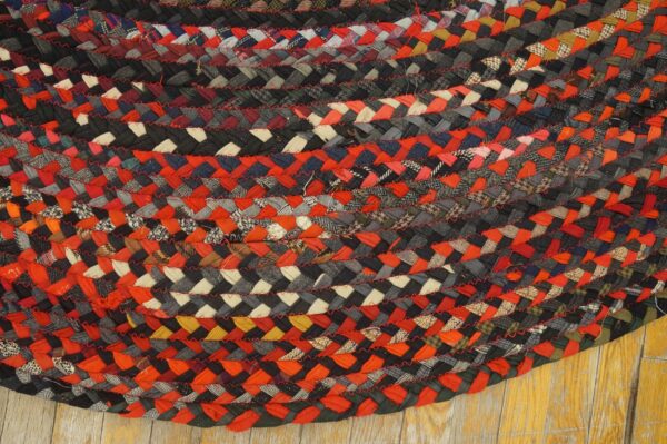 Textured, circular braided rug in black, red, and gray fabric strips with red stitching detail on wood flooring.