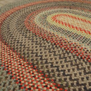 Textured multi-colored braided rug featuring concentric oval rings of red, blue, and earth tones on wood flooring.
