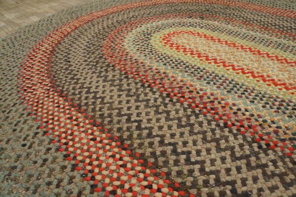 Textured multi-colored braided rug featuring concentric oval rings of red, blue, and earth tones on wood flooring.