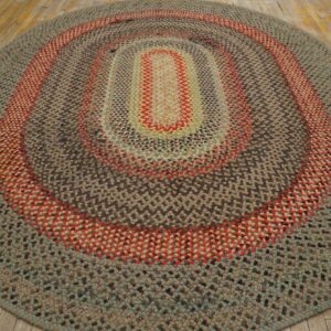 Oval braided rug featuring concentric rings of gray, brown, red, and sage green on rustic light wood flooring.