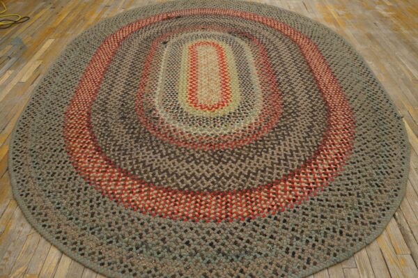 Oval braided rug featuring concentric rings of gray, brown, red, and sage green on rustic light wood flooring.