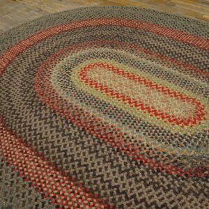 A variegated braided oval rug featuring concentric rings of red, gray, and earthy colors covers rustic wooden planks.