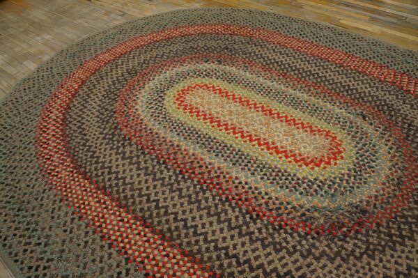 A variegated braided oval rug featuring concentric rings of red, gray, and earthy colors covers rustic wooden planks.