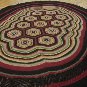 Round rug with black, burgundy, green, and tan crocheted stripes and a central hexagonal pattern.