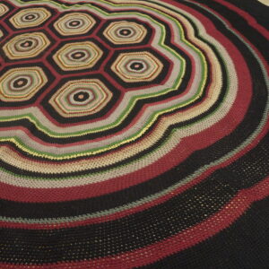 Textured round rug featuring central multicolored hexagons and wide concentric dark stripes on wood flooring.