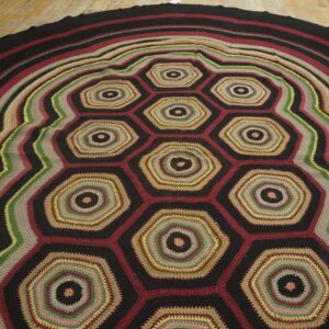 A round, multicolored crochet rug displays an all-over honeycomb pattern and striped border on wooden planks.