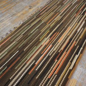 A thick, textured brown rug with multi-colored striped coils rests diagonally on distressed gray flooring.