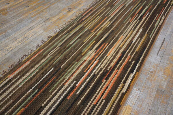 A thick, textured brown rug with multi-colored striped coils rests diagonally on distressed gray flooring.