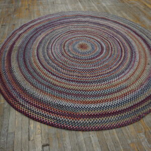 A round, braided rug in navy, red, and cream rests on a rustic, distressed wooden floor.