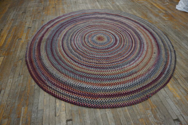 A round, braided rug in navy, red, and cream rests on a rustic, distressed wooden floor.