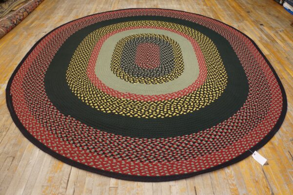 Round braided rug with concentric bands of red, sage, dark green, yellow, and black on wood flooring.