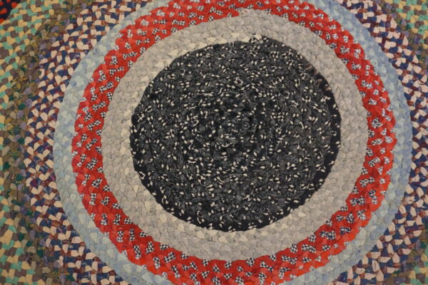 A round braided rug with concentric rings of speckled dark fabric, light gray, patterned red, and light blue.