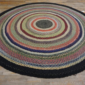 Round braided rug in variegated concentric colors, bordered in black, on a rustic wood floor.
