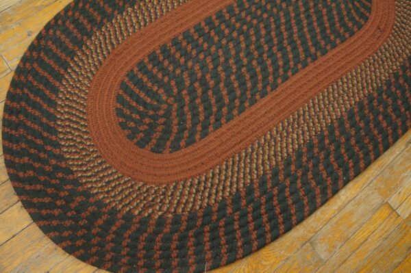 Braided oval rug in rust, black, and tan concentric patterns lying on distressed wood floor.