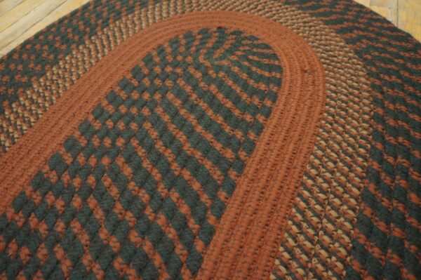 Rust, tan, and dark green braided rug with checkered bands rests on light wooden floorboards.