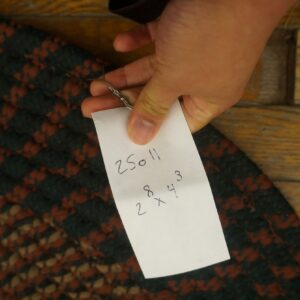 A hand holds a tag over a braided rug featuring dark green, rust, and tan patterns on wood floorboards.