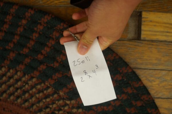 A hand holds a tag over a braided rug featuring dark green, rust, and tan patterns on wood floorboards.